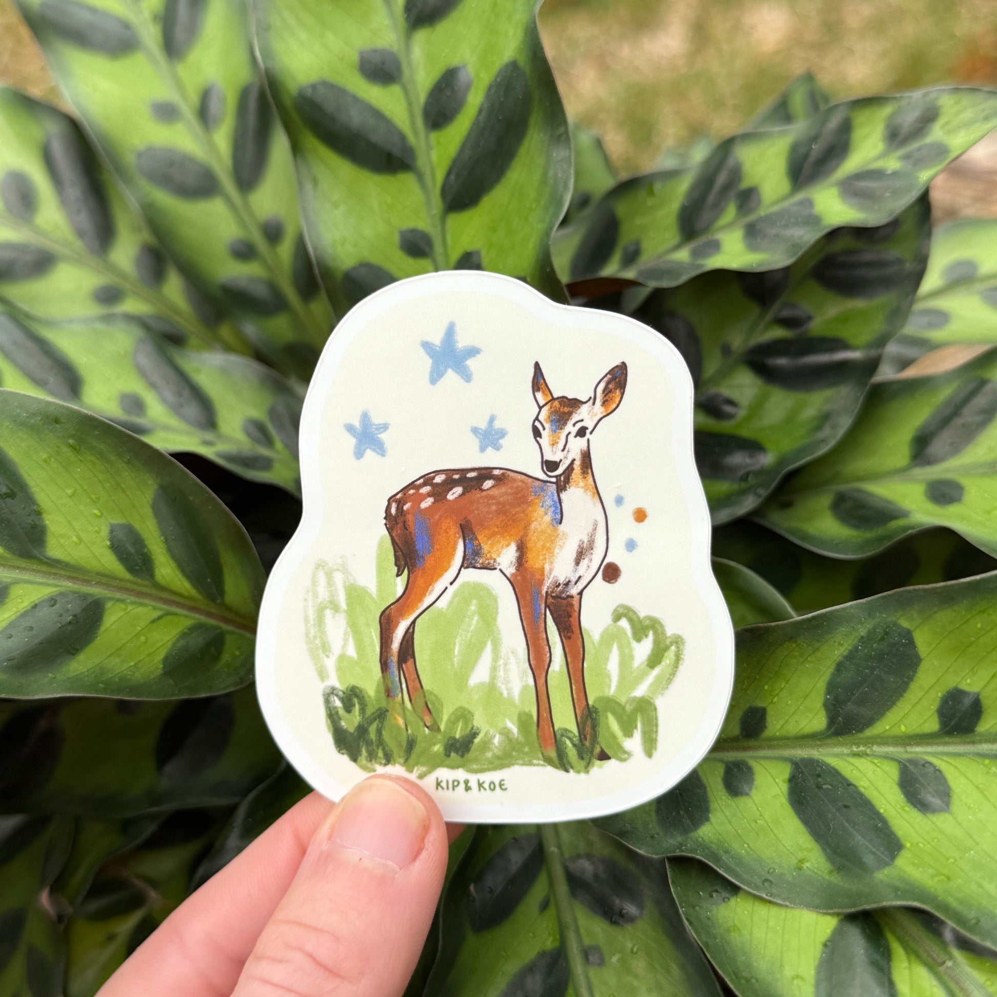 Hand holding a sticker with a deer design against a leafy green background